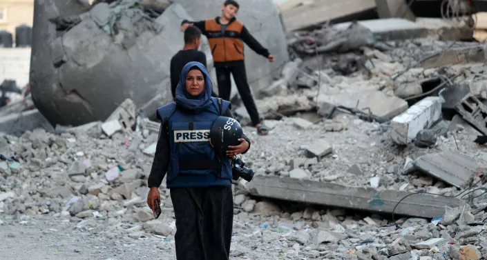 Palestinian journalist walking against backdrop of destruction