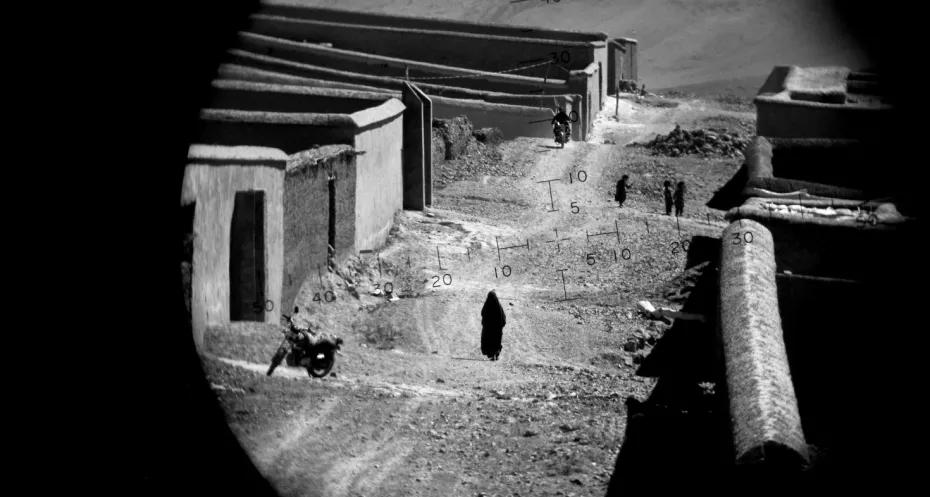 A black and white photograph through the scope of a rifle. The photo shows mud houses with a woman in the distance walking in a burqa. Two children and a motorcyclist approach her.