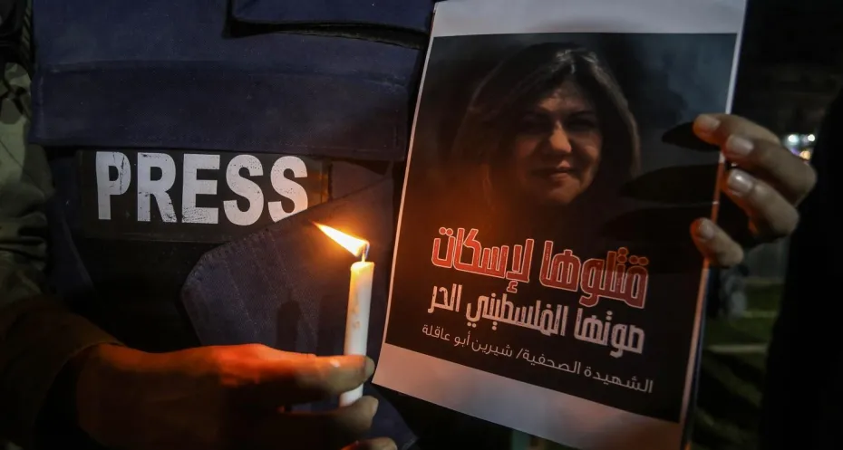 A man in an armored vest written PRESS on it is holding a candle next to the photo of late Palestinian American journalist Shireen Abu Akleh